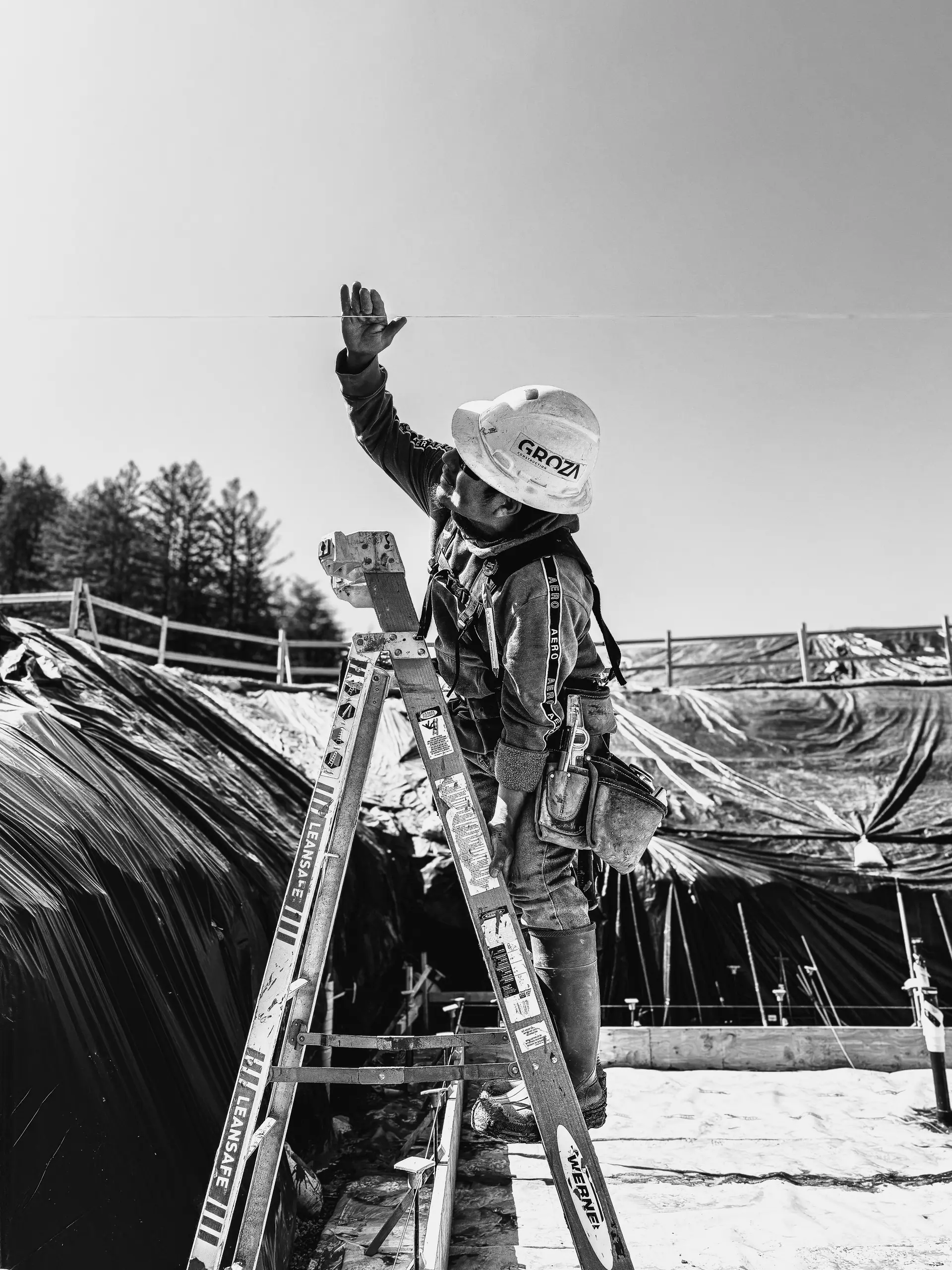 Black-and-white photo of a crew member in a hard hat reaching up from a ladder on site