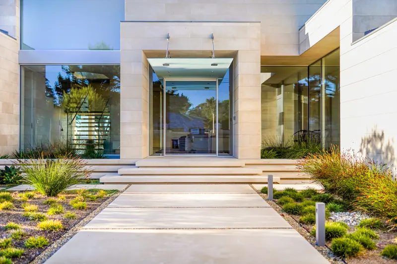 Limestone and glass entry reached by a wide concrete walk through native plantings
