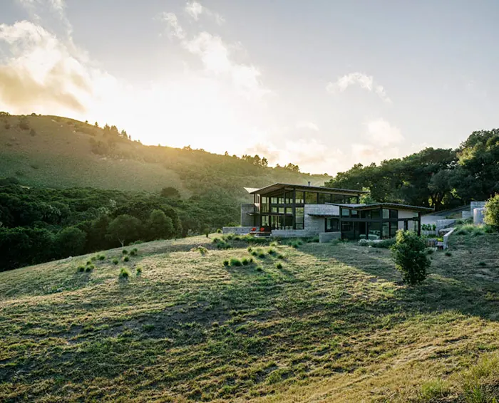 Glass-walled home on a sunlit, grassy hillside at golden hour