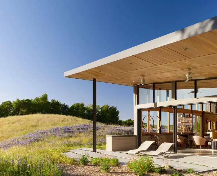 Deep roof overhang shelters an open-air living pavilion above a wildflower meadow