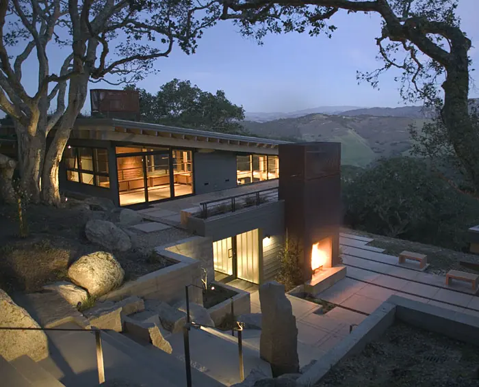 Evening view of a stone and timber home with glowing windows tucked beneath an old oak