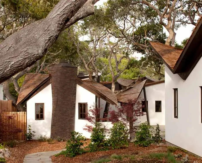 White cottage-style home with steep gables and a dark chimney among old trees