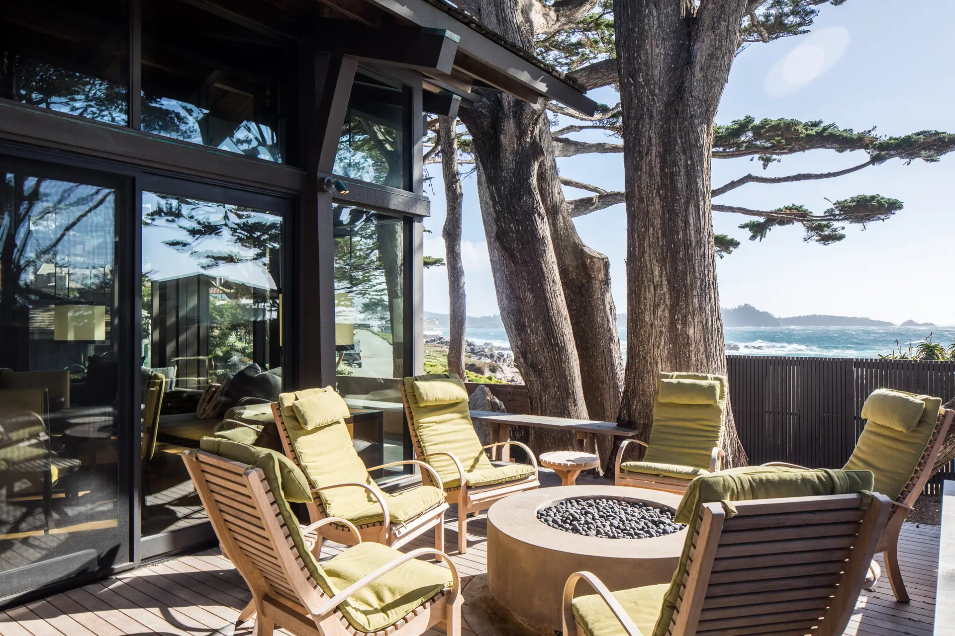 Covered terrace with lounge chairs and a fire bowl under Monterey cypress, ocean beyond