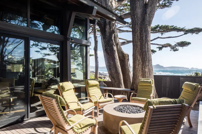 Covered terrace with lounge chairs and a fire bowl under Monterey cypress, ocean beyond