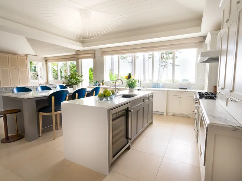Bright white kitchen with a large island and blue stools beneath tall windows
