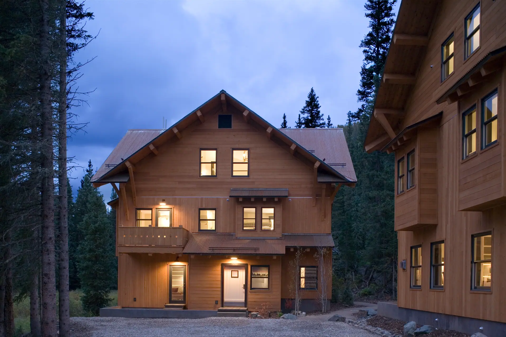 Craftsman-style timber lodge with lit windows at dusk among tall evergreens