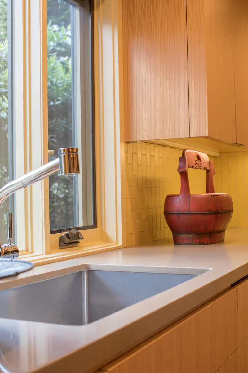 Birch cabinetry, a stainless sink and a red ceramic jug by a sunny window