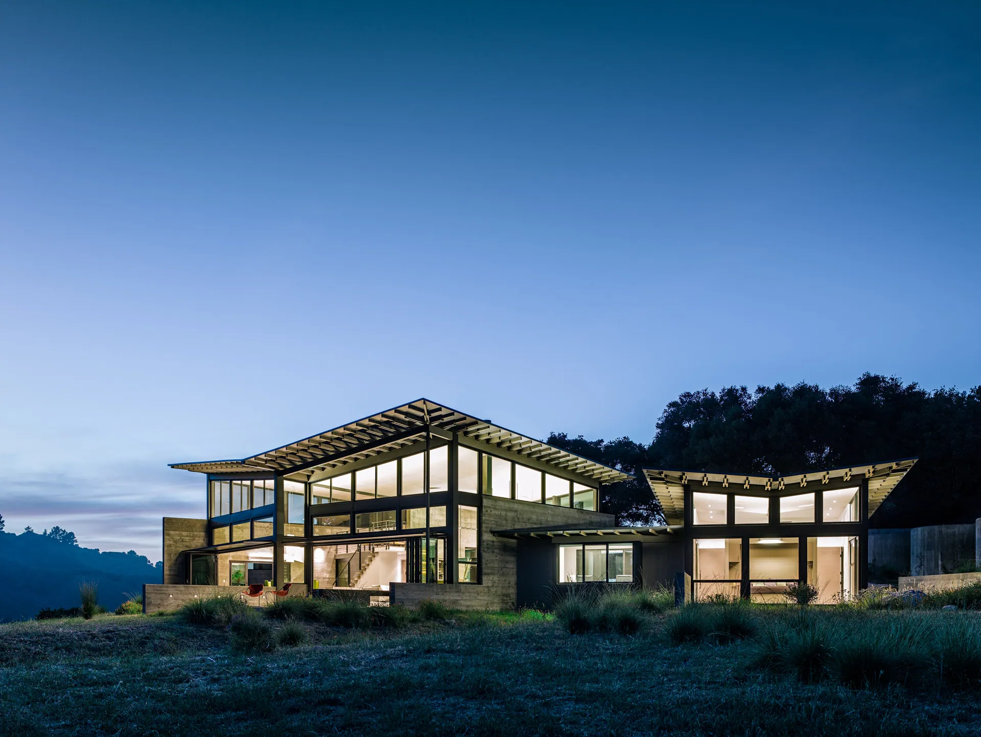 A steel-and-glass home with butterfly roofs glowing at dusk on a Santa Lucia Preserve hillside
