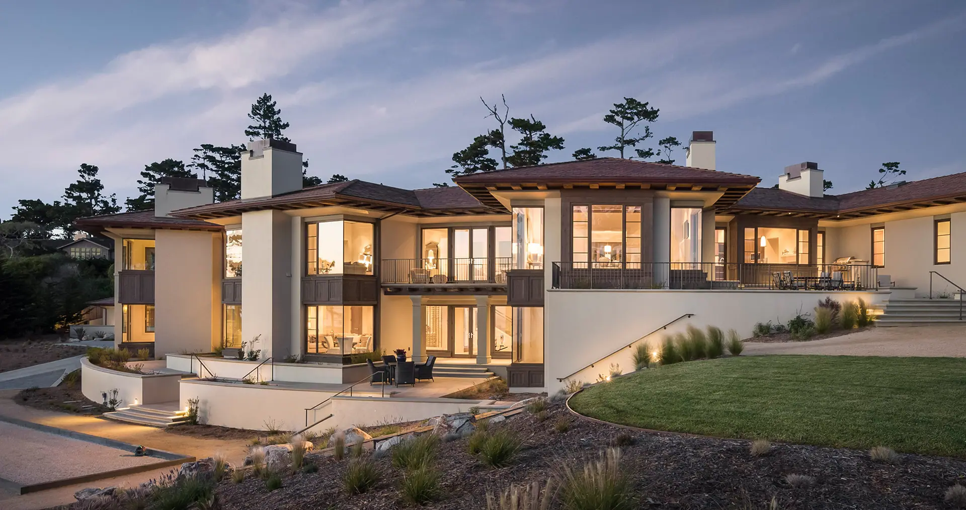 Stucco home with warm lit windows at dusk, a green lawn and Monterey pines behind