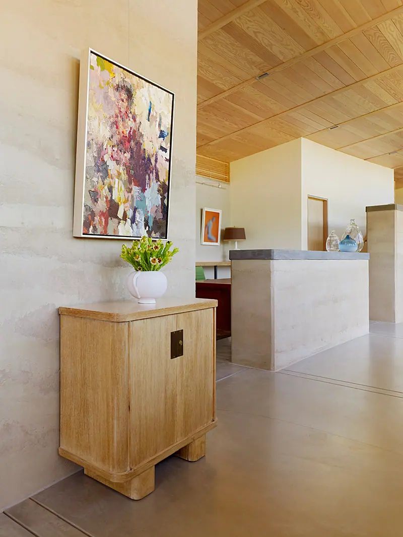 Bold abstract painting above a carved wood cabinet in a rammed-earth hallway